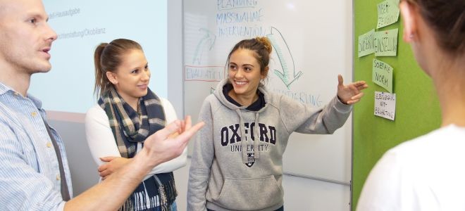 Drei Studierende unterhalten sich fröhlich über eine Pinwand